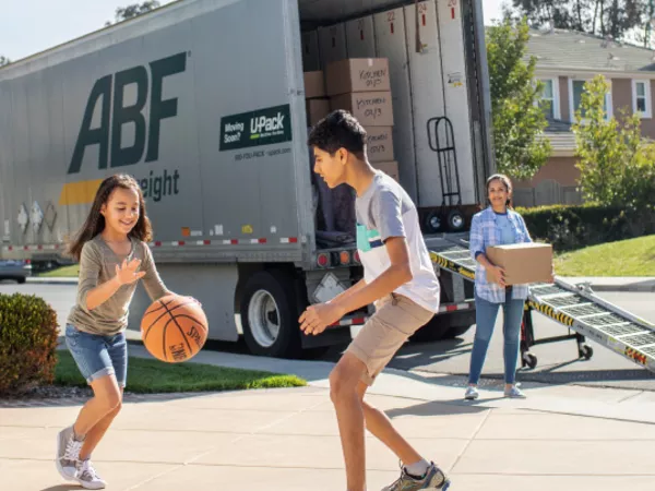Kids play basketball as parents unload a U-Pack moving trailer.