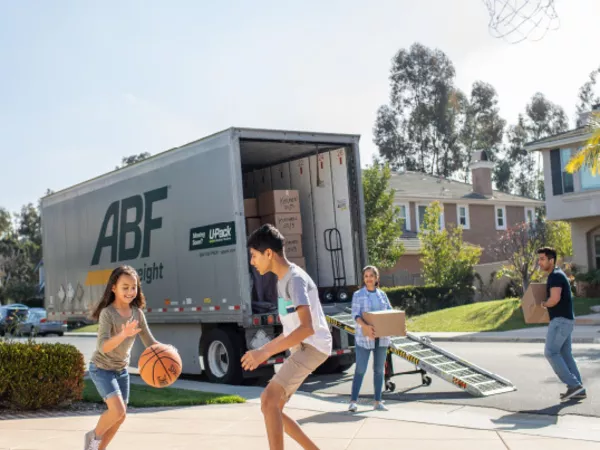 Children play basketball in a driveway while parents load a freight trailer for moving.