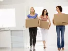 Three women carrying moving boxes, helping their friend move.