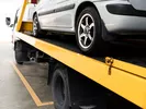 A car transportation company drives a silver car on a yellow auto transport trailer.